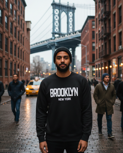 Man wearing a black sweatshirt with 'Brooklyn New York' text, standing in an urban street with the Manhattan Bridge in the background.